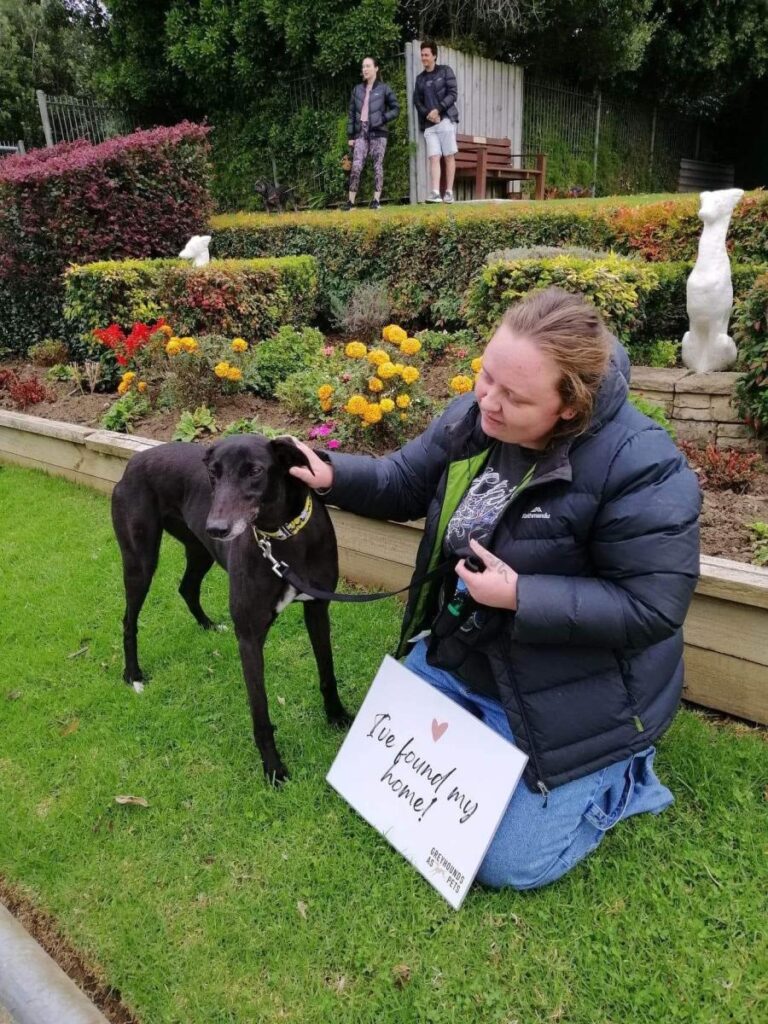 a woman wearing a winter coat in a garden, holding a greyhound's leash. She is kneeling behind a sign that says "I found my home!"